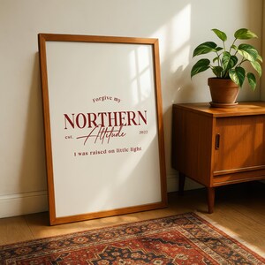 Framed typography print leaning against a wall beside a wooden cabinet and indoor plants on a patterned rug.