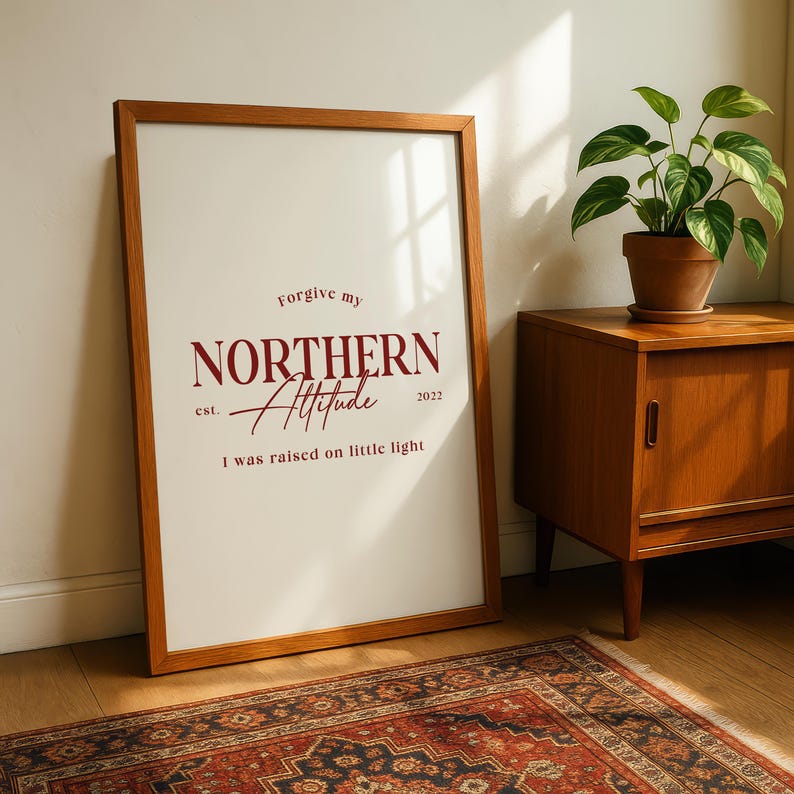 Framed typography print leaning against a wall beside a wooden cabinet and indoor plants on a patterned rug.