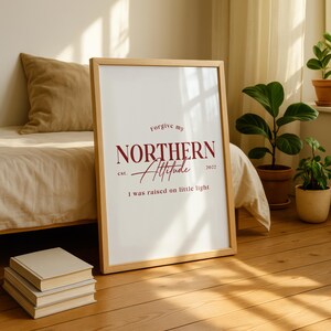 Framed typography print leaning against a wall in a bedroom setting with a bed, pillows, and sunlight coming through a window.
