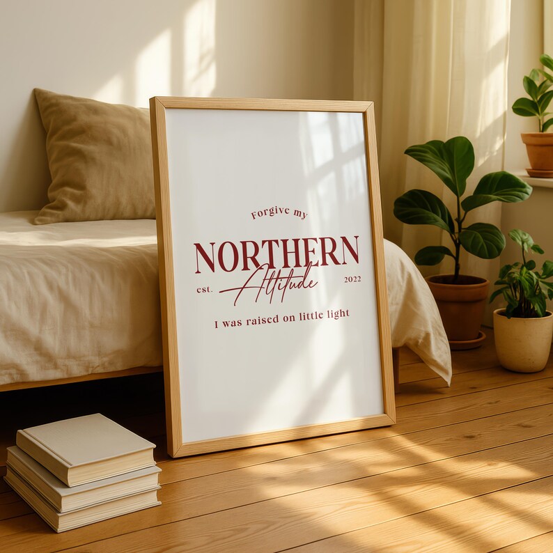 Framed typography print leaning against a wall in a bedroom setting with a bed, pillows, and sunlight coming through a window.