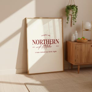 Framed typography print leaning against a wall next to a wooden sideboard with decorative objects and a hanging plant.