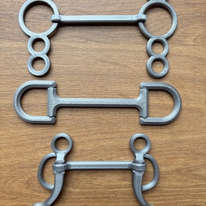 Könnte beinhalten: Drei graue Metallpferdegebisse werden auf einer Holzoberfläche gezeigt. Die Gebisse haben unterschiedliche Designs, darunter ein Ringgebiss, ein D-Ringgebiss und ein Kandarengebiss. Die Gebisse bestehen aus einem soliden, langlebigen Material.