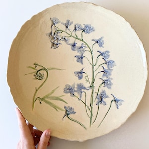 May include: A large, round ceramic plate with a cream-colored base and a scalloped edge. The plate features a hand-painted floral design with blue and green accents. The flowers are delicate and detailed.
