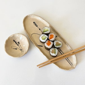 May include: A beige ceramic sushi serving platter and small bowl with black floral designs. The platter holds sushi rolls with avocado and carrot fillings. Bamboo chopsticks rest on the platter. The items are on a white surface.