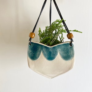 May include: A white ceramic hanging planter with a blue and white swirl design. The planter has a scalloped edge and is hanging from a black leather cord with wooden beads.