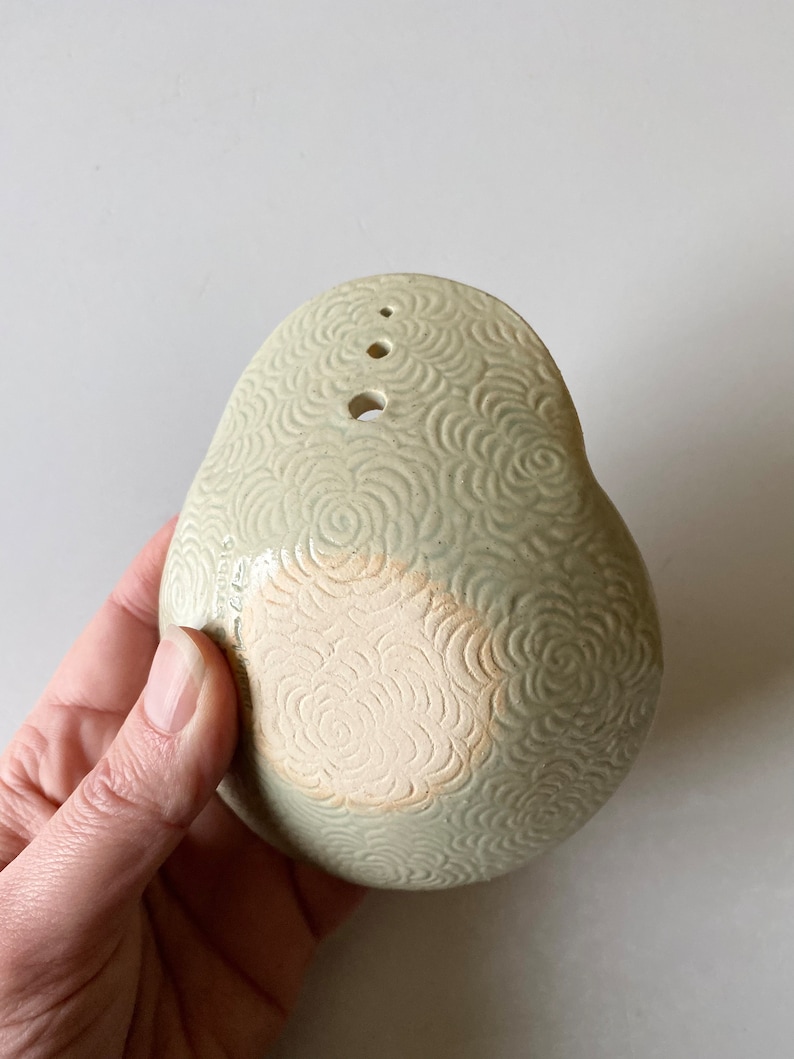 May include: A light green ceramic bowl with a textured floral pattern. The bowl has four small holes on the top and a larger hole on the bottom.