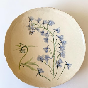 May include: A round, off-white ceramic plate with a scalloped edge. Delicate blue floral artwork adorns the plate, featuring a detailed botanical design of blue flowers and green leaves. The plate has a rustic, handmade aesthetic.