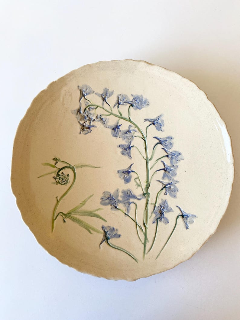 May include: A round, off-white ceramic plate with a scalloped edge. Delicate blue floral artwork adorns the plate, featuring a detailed botanical design of blue flowers and green leaves. The plate has a rustic, handmade aesthetic.