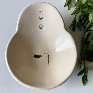 May include: A white ceramic dish with a small black plant design and three holes on the top. The dish is slightly oval shaped.