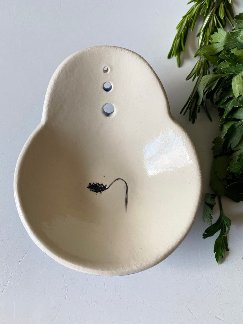 May include: A white ceramic dish with a small black plant design and three holes on the top. The dish is slightly oval shaped.