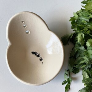 May include: A white ceramic bowl with a small black floral design. The bowl has three small holes on the side and a slightly irregular shape.