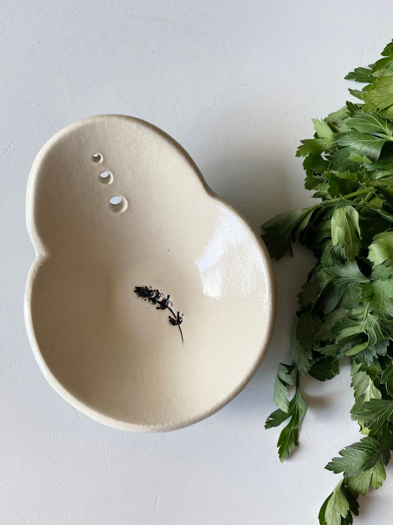 May include: A white ceramic bowl with a small black floral design. The bowl has three small holes on the side and a slightly irregular shape.
