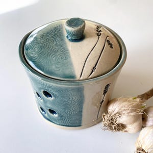 May include: A ceramic garlic keeper with a lid. The keeper is divided into two sections, one teal with a swirl pattern and the other off-white with a floral design. The lid has a small knob and the same color scheme. Garlic bulbs are next to the keeper.