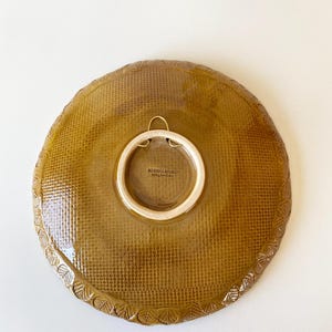 May include: A round, ceramic plate with a textured, woven pattern in a warm, golden-brown hue. The plate features a decorative, leaf-like border and a white, circular attachment for hanging. The back of the plate has the text "BOTANICA STUDIO".