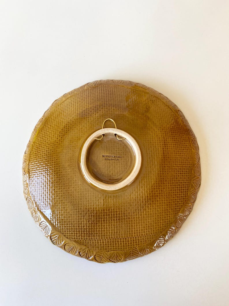May include: A round, ceramic plate with a textured, woven pattern in a warm, golden-brown hue. The plate features a decorative, leaf-like border and a white, circular attachment for hanging. The back of the plate has the text "BOTANICA STUDIO".