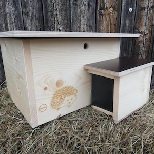 Könnte beinhalten: Zwei Igelhäuser aus Holz, eines größer als das andere, mit einer kleinen Öffnung oben auf dem größeren Haus. Die Häuser stehen auf einem Strohboden.