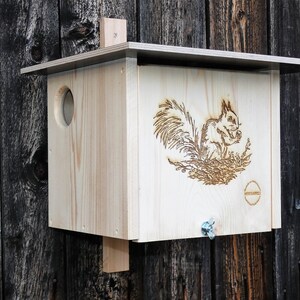 Könnte beinhalten: Ein Vogelhaus aus Holz mit einem Eichhörnchen-Motiv auf der Vorderseite. Das Vogelhaus hat eine kleine Öffnung für Vögel, um einzudringen, und ein flaches Dach.