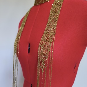 Könnte beinhalten: Ein goldfarbenes, mit Perlen besetztes Schal- und Choker-Set. Der Choker liegt eng am Hals an, und der Schal hängt nach vorne. Das Set wird auf einer roten Schneiderpuppe präsentiert. Der Schal besteht aus kleinen, glänzenden Perlen.