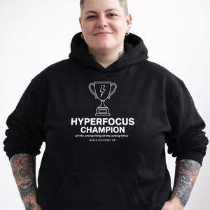 Puede incluir: Sudadera con capucha negra con un bolsillo frontal y un gráfico blanco. El gráfico presenta un trofeo con un rayo y el texto "HYPERFOCUS CHAMPION (of the wrong thing at the wrong time) WIRED DIFFERENT CO."