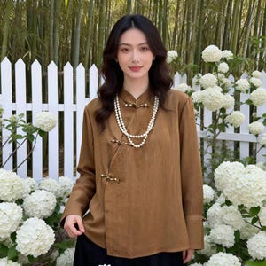 May include: A brown long-sleeve shirt with a mandarin collar and side closures. The shirt is paired with a black skirt and a pearl necklace. The background features a white picket fence and white flowers.