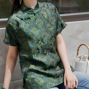 Puede incluir: Top verde con estampado floral, mangas cortas y cierres de rana. El top presenta un cuello mandarín y un patrón de flores azules y verdes. El top se combina con unos vaqueros azul claro.