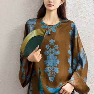 May include: A brown long-sleeved blouse featuring a vibrant blue floral and bird pattern, paired with a handheld folding fan that has a dark green surface and a matching green tassel hanging from the handle, worn with light blue denim jeans.