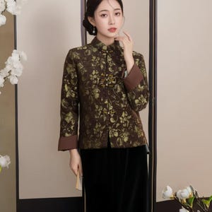 Puede incluir: Una mujer con una chaqueta marrón con estampado floral, cuello mandarín y cierres de rana dorados. La chaqueta tiene puños marrones y se combina con una falda negra. El fondo incluye una biombo y flores blancas.
