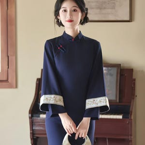 May include: A navy blue, long-sleeved dress with a mandarin collar and floral embroidery on the cuffs. The dress features a side slit and is paired with a cream-colored handbag. The woman is standing in front of a piano.