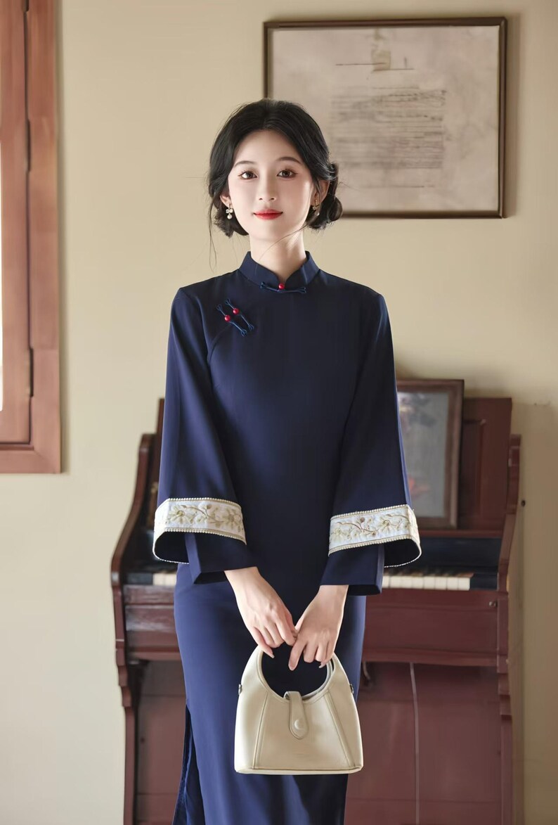 May include: A navy blue, long-sleeved dress with a mandarin collar and floral embroidery on the cuffs. The dress features a side slit and is paired with a cream-colored handbag. The woman is standing in front of a piano.