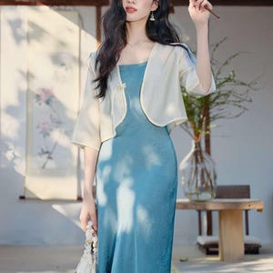 May include: A woman wearing a light blue dress with a matching cream-colored bolero jacket. She is holding a fan and a small white purse. The dress has a square neckline and a subtle floral pattern.