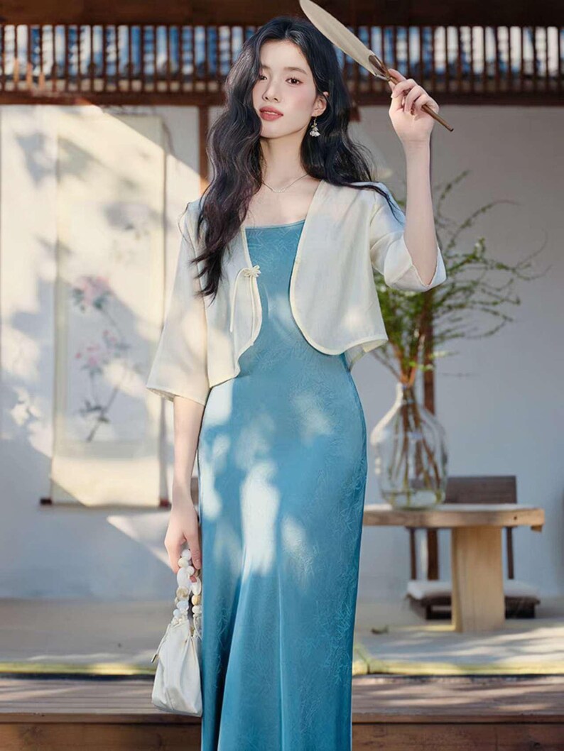 May include: A woman wearing a light blue dress with a matching cream-colored bolero jacket. She is holding a fan and a small white purse. The dress has a square neckline and a subtle floral pattern.
