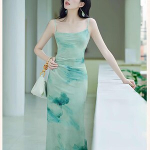 Puede incluir: Un vestido largo verde claro con un dise&ntilde;o similar a una acuarela. El vestido presenta finos tirantes finos y un corpi&ntilde;o ajustado que fluye hacia una falda ligeramente acampanada. El dise&ntilde;o incluye tonos de azul y verde, creando un aspecto suave y et&eacute;reo. Tambi&eacute;n se ve un peque&ntilde;o bolso blanco con cuentas de madera.