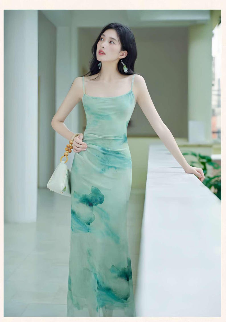 Puede incluir: Un vestido largo verde claro con un dise&ntilde;o similar a una acuarela. El vestido presenta finos tirantes finos y un corpi&ntilde;o ajustado que fluye hacia una falda ligeramente acampanada. El dise&ntilde;o incluye tonos de azul y verde, creando un aspecto suave y et&eacute;reo. Tambi&eacute;n se ve un peque&ntilde;o bolso blanco con cuentas de madera.