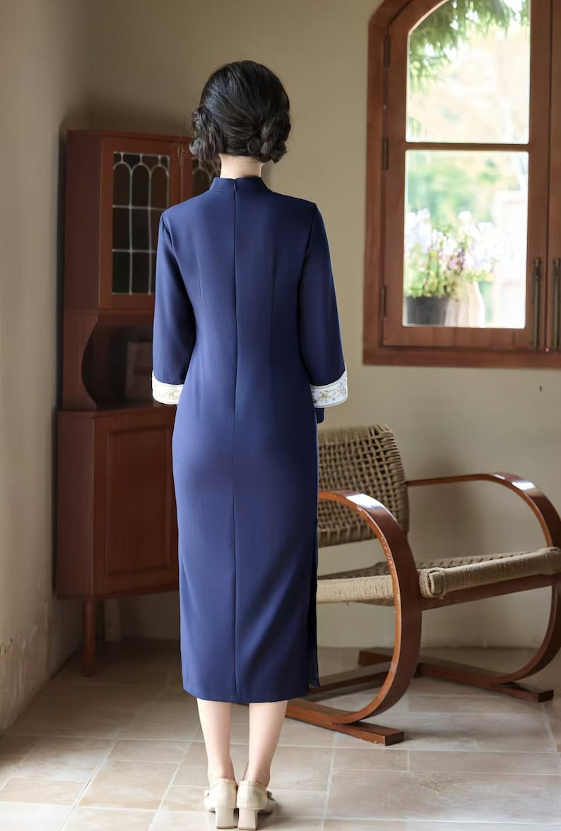 May include: A navy blue, long-sleeve dress with a mandarin collar and a side slit. The cuffs feature white and gold embroidered detailing. The dress is knee-length and has a slim fit. The background includes a wooden cabinet, a chair, and a window.