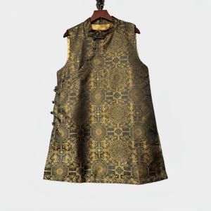 May include: A gold and black patterned vest, sleeveless with a mandarin collar and decorative buttons. The fabric has a repeating geometric design. The vest is hanging on a wooden hanger.