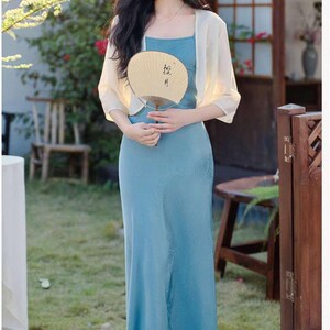 May include: A woman wearing a light blue dress with a square neckline and a matching sheer, cream-colored shawl. She is holding a beige hand fan with black calligraphy. The scene is outdoors, with greenery and a wooden door in the background.