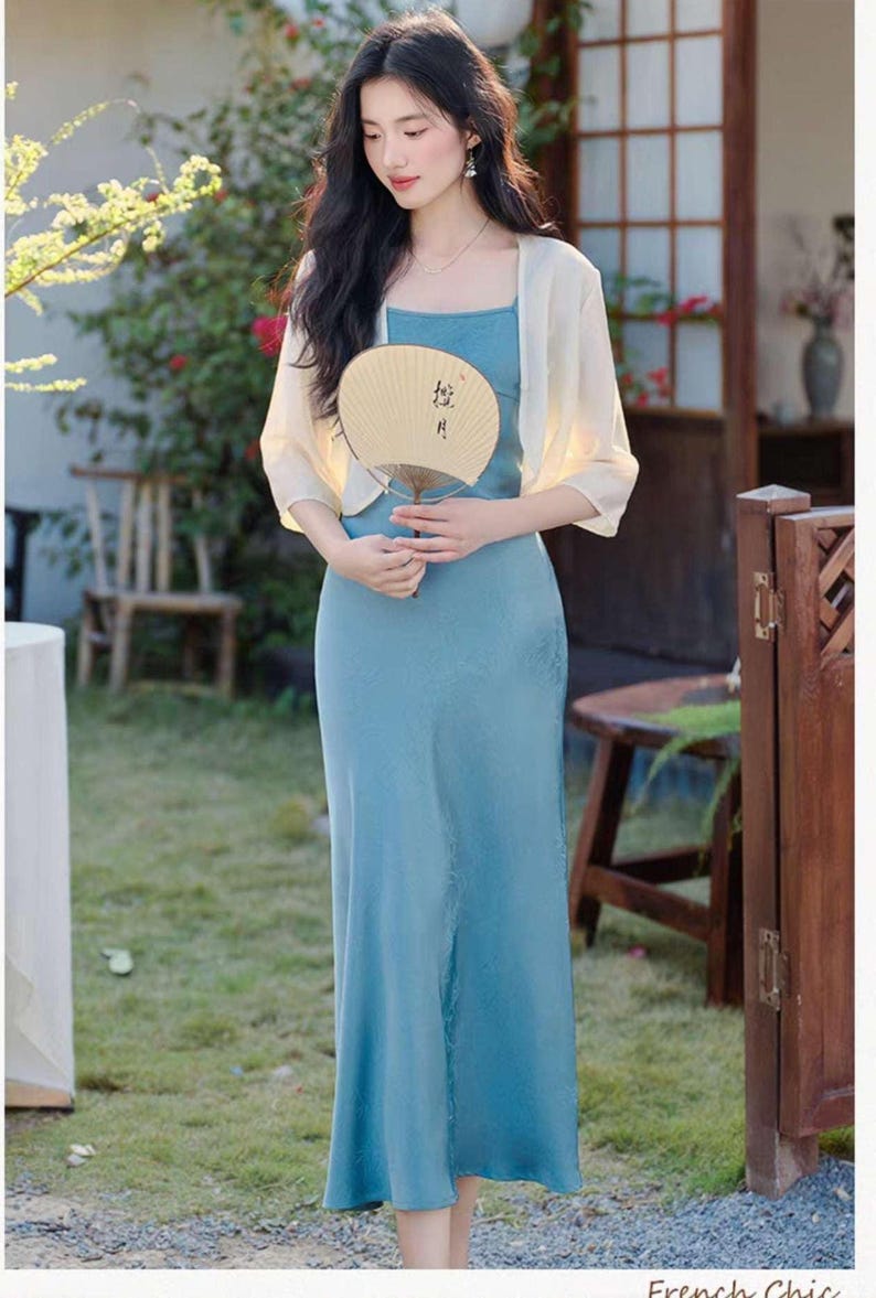 May include: A woman wearing a light blue dress with a square neckline and a matching sheer, cream-colored shawl. She is holding a beige hand fan with black calligraphy. The scene is outdoors, with greenery and a wooden door in the background.