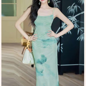 Puede incluir: Un vestido largo verde claro con tirantes finos y un estampado tipo acuarela. El vestido se combina con un bolso blanco con asa dorada. El fondo incluye una pantalla negra con un dise&ntilde;o de bamb&uacute;.