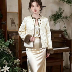 May include: Cream-colored satin ensemble featuring a long skirt and a matching jacket with floral embroidery. The jacket has brown fur cuffs and trim. A long beaded necklace and a cream-colored camisole complete the outfit. The outfit is suitable for formal occasions.