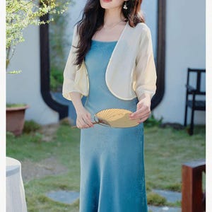 May include: A woman wearing a light blue dress and a cream-colored bolero jacket, holding a gold fan. The dress has a floral pattern. The background includes greenery and a decorative mirror.