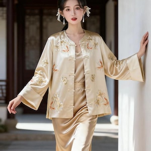 May include: A golden-beige silk pajama set featuring a jacket with floral embroidery and wide sleeves, a matching camisole, and a long skirt. The jacket has a mandarin collar and frog closures. The embroidery includes orange and white floral designs.