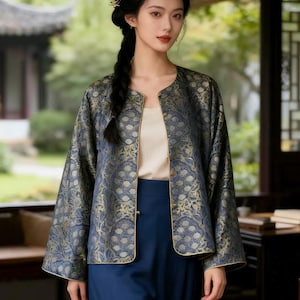May include: A woman wearing a traditional Chinese-style outfit. The outfit includes a navy blue jacket with gold floral patterns, a cream-colored top, and navy blue pants. The jacket has gold trim and the outfit is worn outdoors.