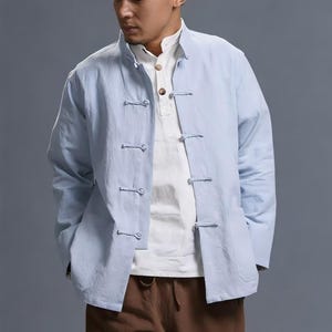 Peut inclure: Veste bleu clair avec col officier et fermetures à brandebourgs, portée sur une chemise blanche. La veste a des manches longues et deux poches avant. La personne porte également un pantalon marron.