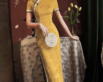 Puede incluir: Vestido amarillo mostaza de manga corta, cuello alto y abertura lateral. El vestido presenta ribetes negros y detalles de encaje. Se sostiene en la mano un pequeño bolso de mano circular blanco. El vestido es de estilo tradicional.