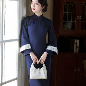 May include: A navy blue, long-sleeve dress with a mandarin collar and flared sleeves. The cuffs feature white and gold embroidery. A white handbag is held in front of the dress. The dress has a side slit.