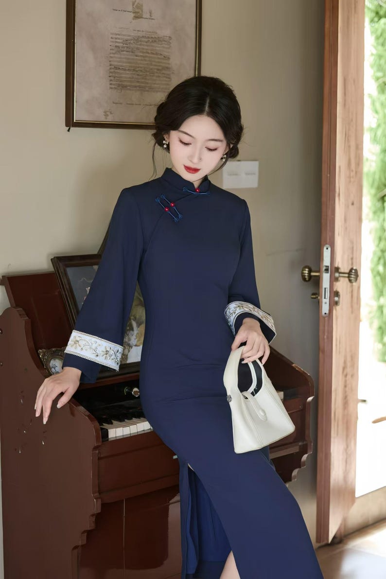 May include: A navy blue, long-sleeved dress with a mandarin collar and a side slit. The dress features embroidered cuffs with floral patterns and decorative buttons. A white handbag is held in the hand.