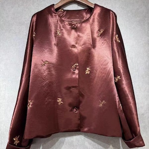 May include: A maroon satin blouse with long sleeves and a round neckline. The blouse features gold floral embroidery and a button-down front. The fabric has a subtle sheen, and the garment is hanging on a wooden hanger.