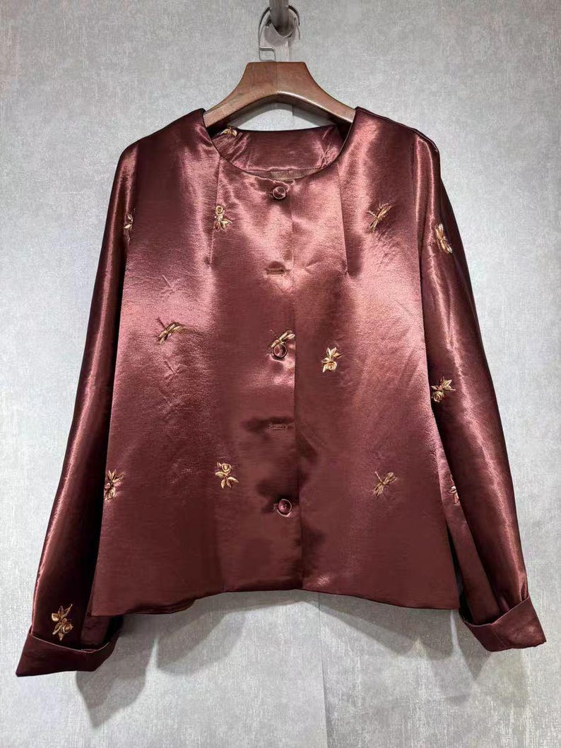 May include: A maroon satin blouse with long sleeves and a round neckline. The blouse features gold floral embroidery and a button-down front. The fabric has a subtle sheen, and the garment is hanging on a wooden hanger.