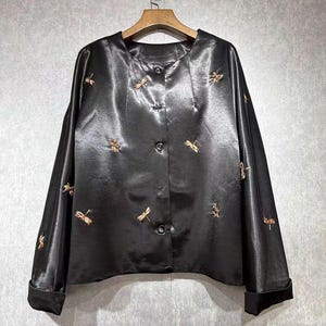 May include: Black satin button-down blouse with long sleeves and gold dragonfly embroidery. The blouse is hanging on a wooden hanger against a neutral background. The cuffs are folded up.