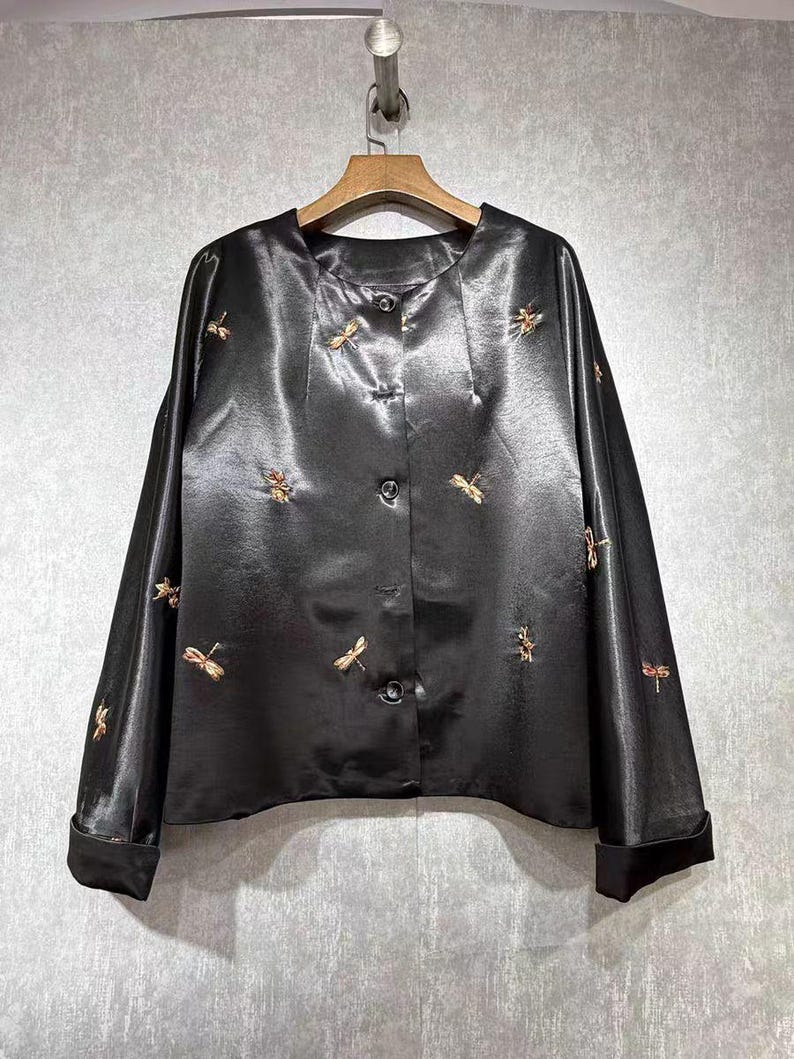 May include: Black satin button-down blouse with long sleeves and gold dragonfly embroidery. The blouse is hanging on a wooden hanger against a neutral background. The cuffs are folded up.